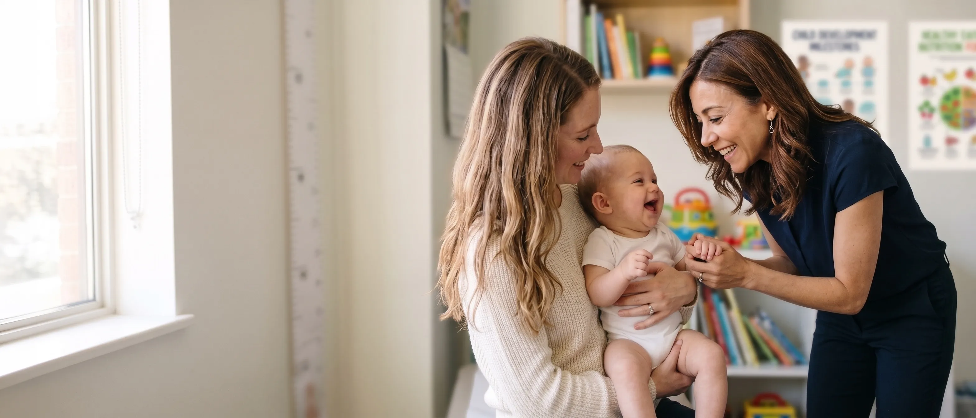 Dr. Erica Bailen leans in to greet a laughing baby held by their mother in an exam room.